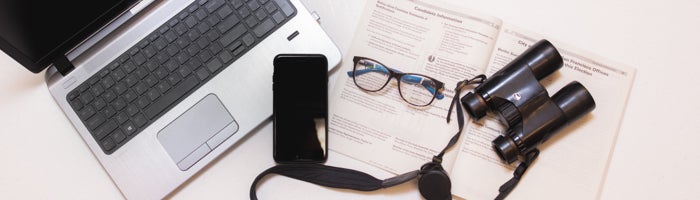 Laptop, mobile phone, binoculars and glasses, and Voter Information Pamphlet displayed on a desk 